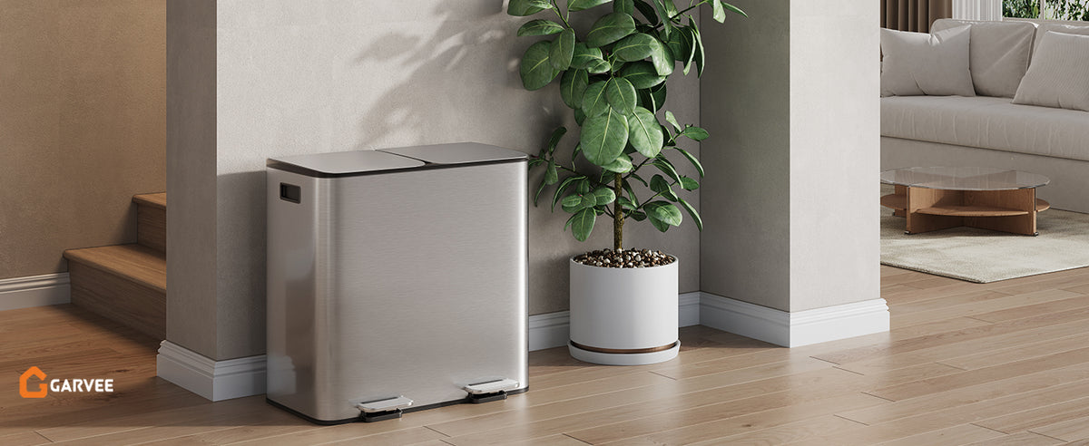 A stainless steel dual compartment trash can and recycling bin placed next to a large potted plant in a modern interior.
