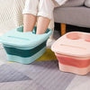 Two collapsible foot soak buckets in teal and pink resting on a gray rug next to a person's legs.
