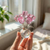 A hand holds a crystal rose flower figurine with pink glass petals in a clear glass vase.