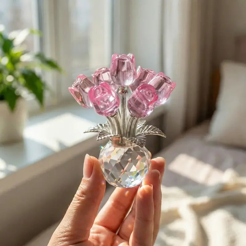 A hand holds a crystal rose flower figurine with pink glass petals in a clear glass vase.