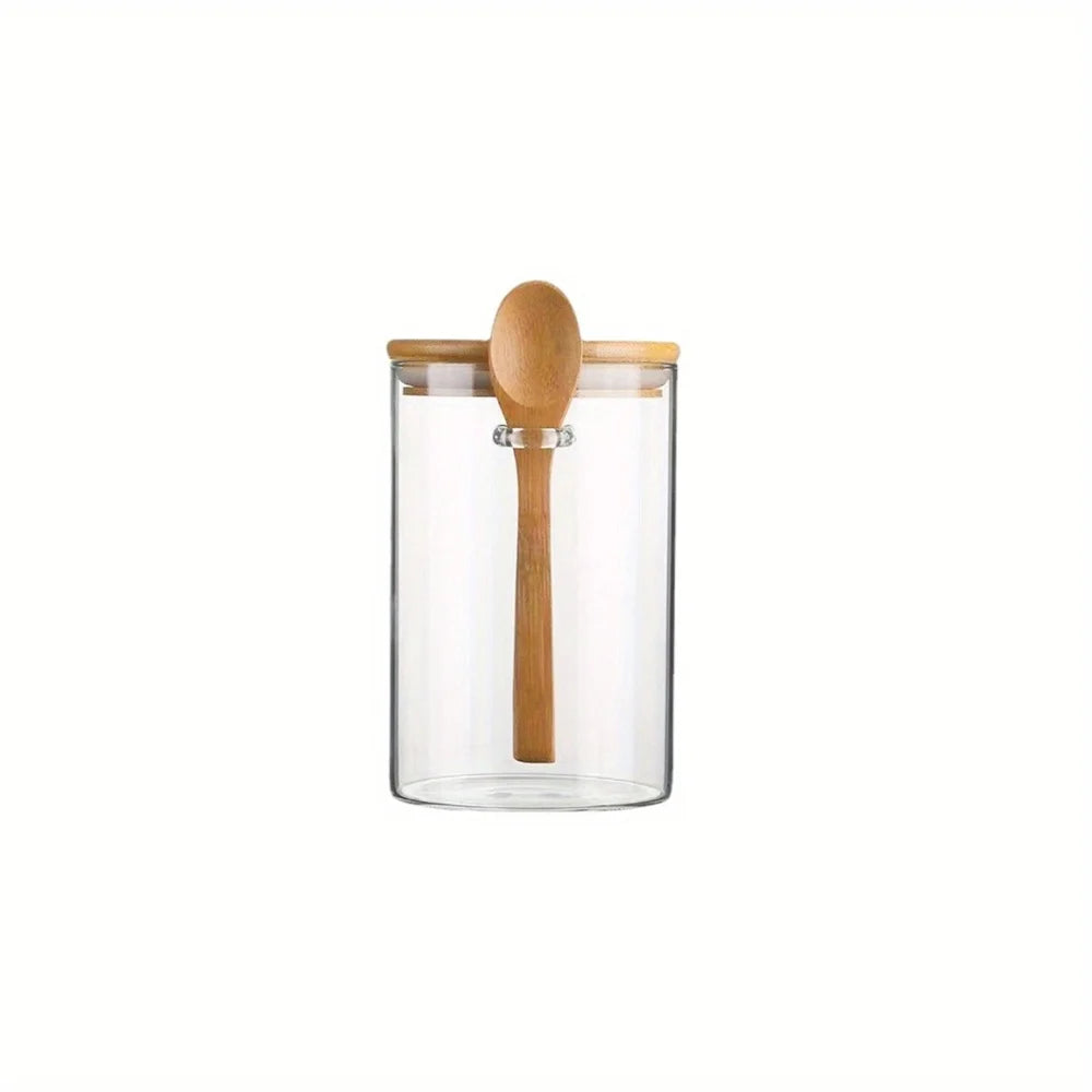 A clear glass spice jar with a bamboo lid and a wooden spoon resting inside.