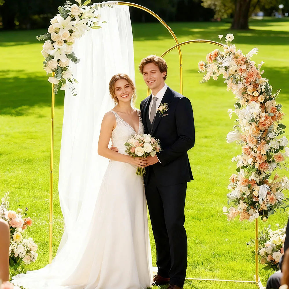 Una novia y un novio están frente a un arco nupcial de metal dorado decorado con flores blancas y melocotón sobre un césped v