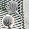 A silver metal cleaning broom with a dense, fluffy head is mounted on a modern window blind.