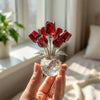 A hand holds a crystal rose flower figurine with red petals in a clear glass vase.