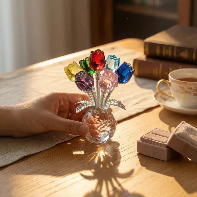 A hand holds a crystal rose flower figurine in a glass vase with colorful petals on a wooden table.