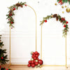 Soporte de arco nupcial de metal dorado decorado con ramas perennes y adornos rojos contra una pared blanca.