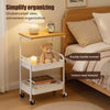 A white 3-tier rolling storage cart with a wooden top, holding a lamp and a teddy bear, next to a wooden bed frame.