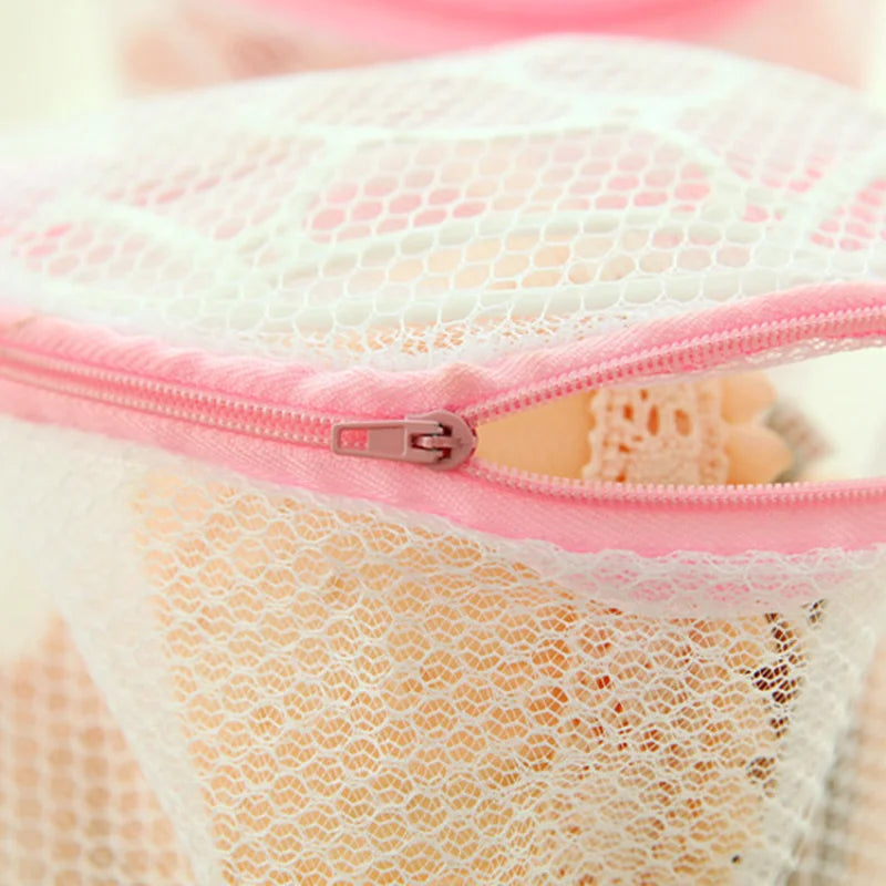 A pink reusable mesh lingerie washing bag with a zipper is shown against a white lace background.