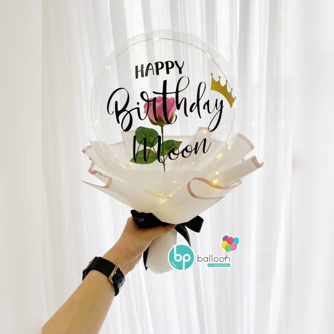 A hand holds a clear latex balloon shaped like a moon with 'Happy Birthday Moon' text and a gold crown.