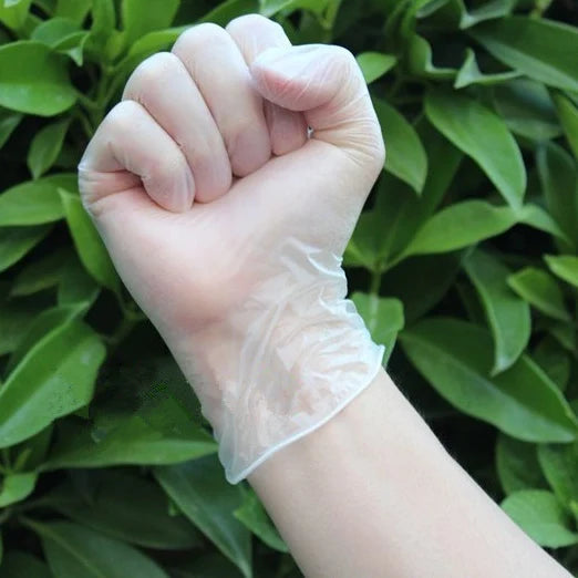 A hand wearing a disposable vinyl cleaning glove is clenched in a fist against a background of green leaves.