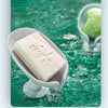A bar of white soap is dripping into clear water, resting in a light gray dish amidst green, textured water.
