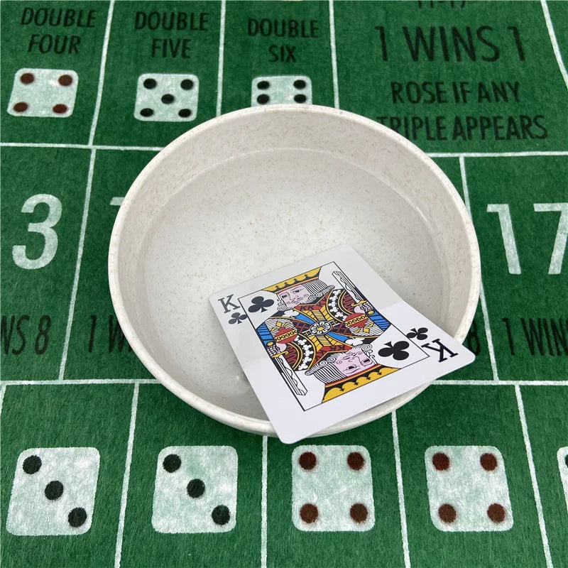 A white bowl holds a playing card on a green felt table with scattered dice and card markings.