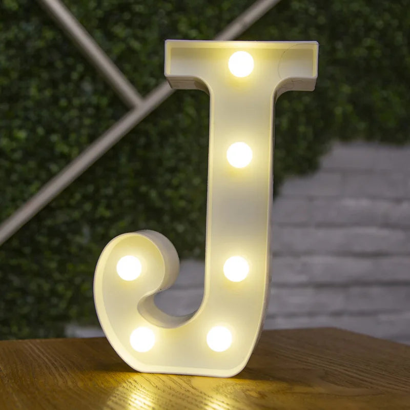 A white letter J made of LED lights standing on a wooden surface with a blurred green background.