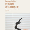 A hand holds a black 3D printed profile eyeglass holder against a white slatted background.