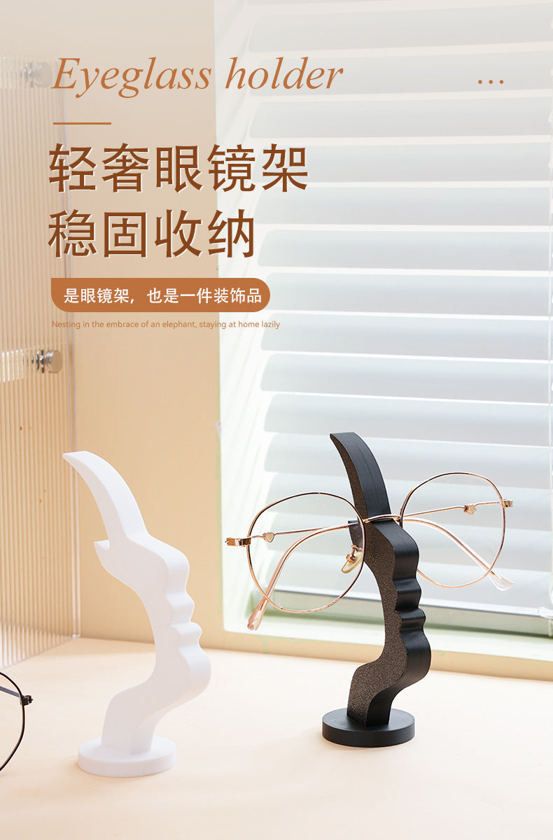 A minimalist black 3D printed profile stand for eyeglasses, shown next to a white stand and a window.