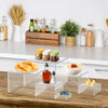 Clear acrylic stackable riser displaying various food items like fruit, nuts, and dips on a wooden surface.