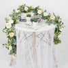 A white table decorated with an artificial eucalyptus and rose garland featuring greenery, roses, and a small potted succulen