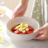 A hand holds a white ceramic soup bowl filled with sliced strawberries and a yellow topping on a white surface.
