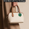 A hand is holding a beige foldable handbag with a green flower charm against a neutral background.