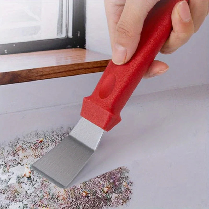 Una mano sostiene un cepillo de cocina rojo con una hoja plateada, apoyado sobre una superficie con residuo de limpieza.