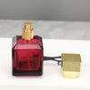 A glass perfume bottle with a red liquid and a gold cap next to a square gold metal dispenser.