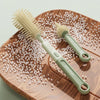 A light green silicone bottle brush with a handle rests on a wooden board surrounded by small white beads.