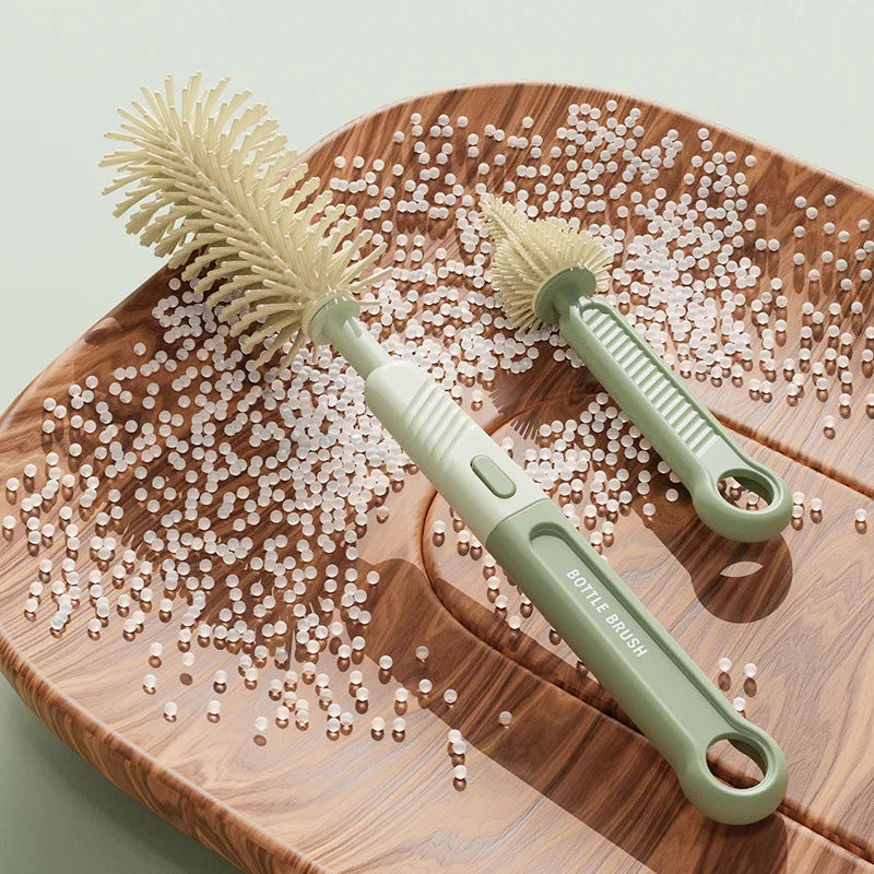 A light green silicone bottle brush with a handle rests on a wooden board surrounded by small white beads.