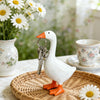 A white goose sculpture with an orange beak and feet is displayed on a woven tray next to a teacup.