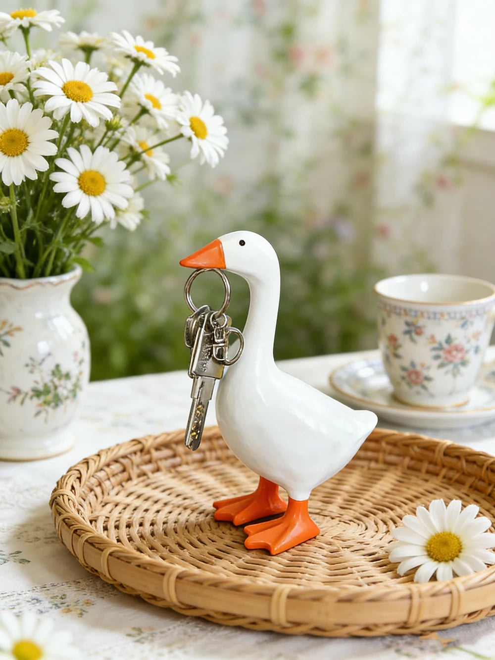 A white goose sculpture with an orange beak and feet is displayed on a woven tray next to a teacup.