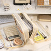 A beige pencil pouch with a transparent lining and various writing tools is displayed on a white shelf.