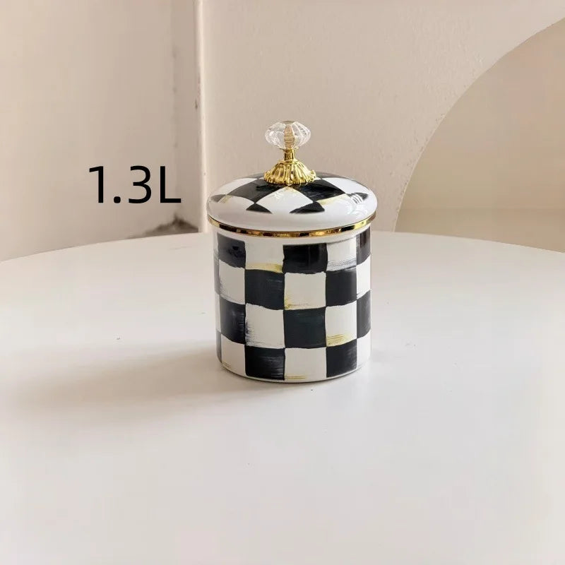 A small storage jar with a checkered black and white pattern and a gold lid sits on a white surface.