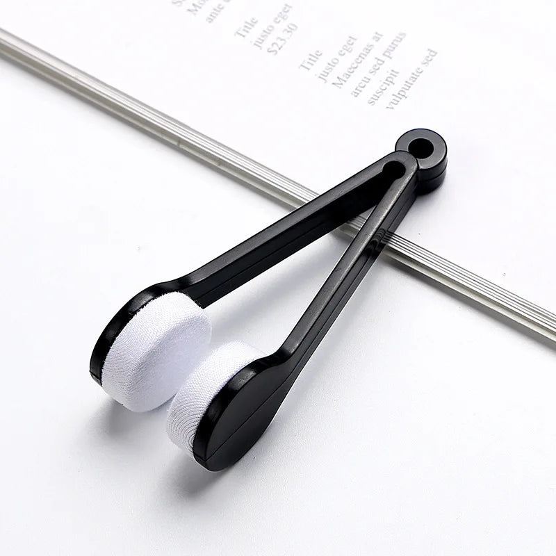 A black silicone cleaning tool with white pads for cleaning eyeglasses is shown on a white background.
