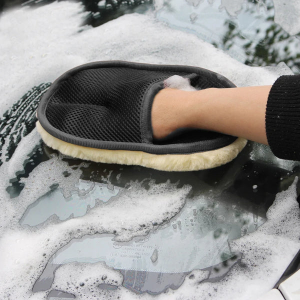 A hand wearing a black soft car washing glove is shown cleaning a car surface covered in soap foam.