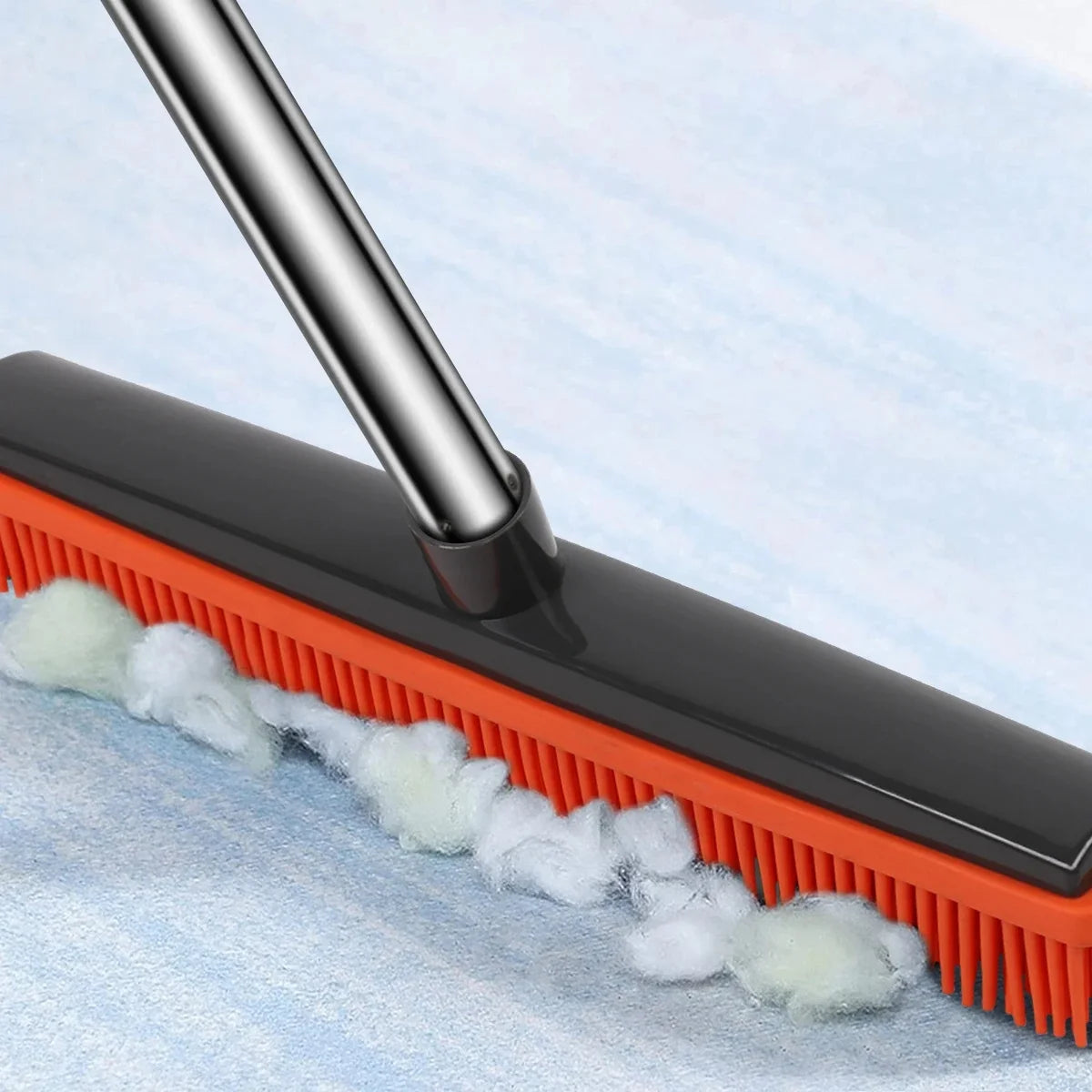 A black rubber broom with orange bristles and a metal squeegee is shown cleaning pet hair from a light blue surface.