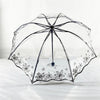 A clear, retro transparent folding umbrella with a black frame and floral pattern rests on white bedding.