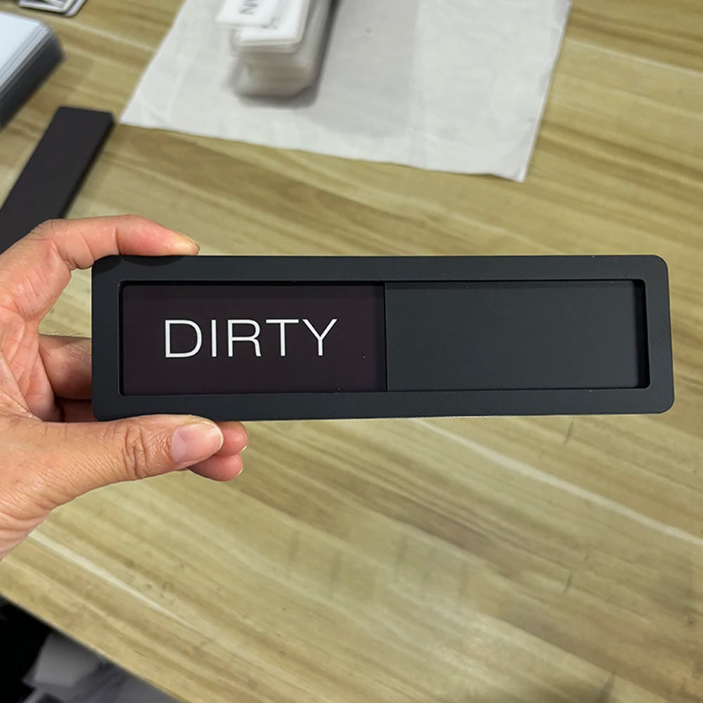 A hand holds a black rectangular magnetic item labeled DIRTY on a wooden surface.