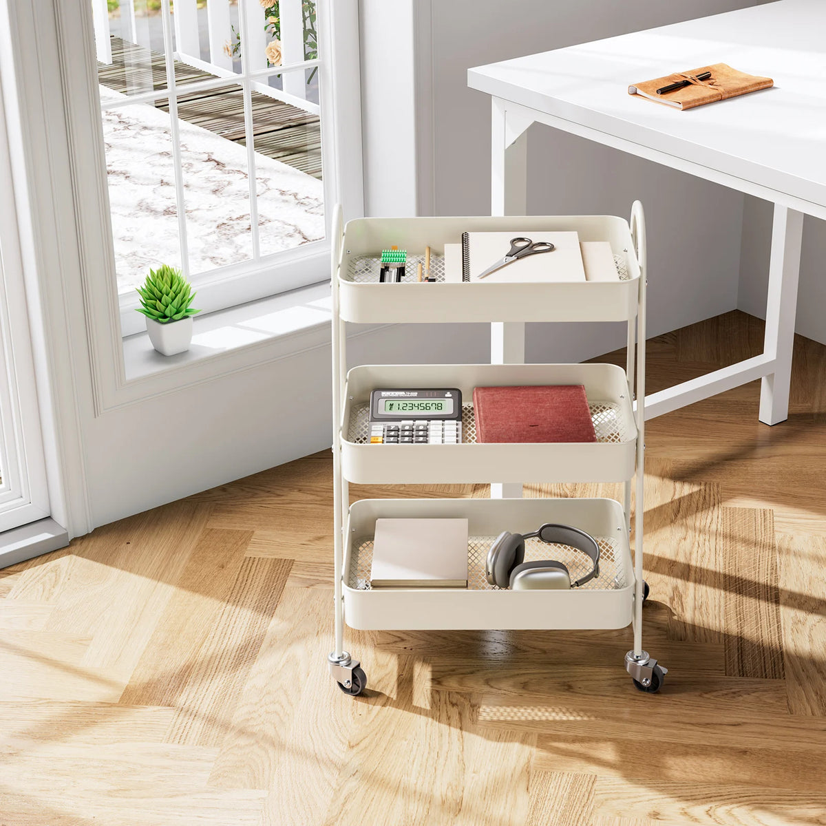 A white metal rolling storage cart with three tiers holds office supplies and electronics on a wooden floor.