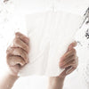 Hands holding a white thick disposable facial cleansing towel under running water.
