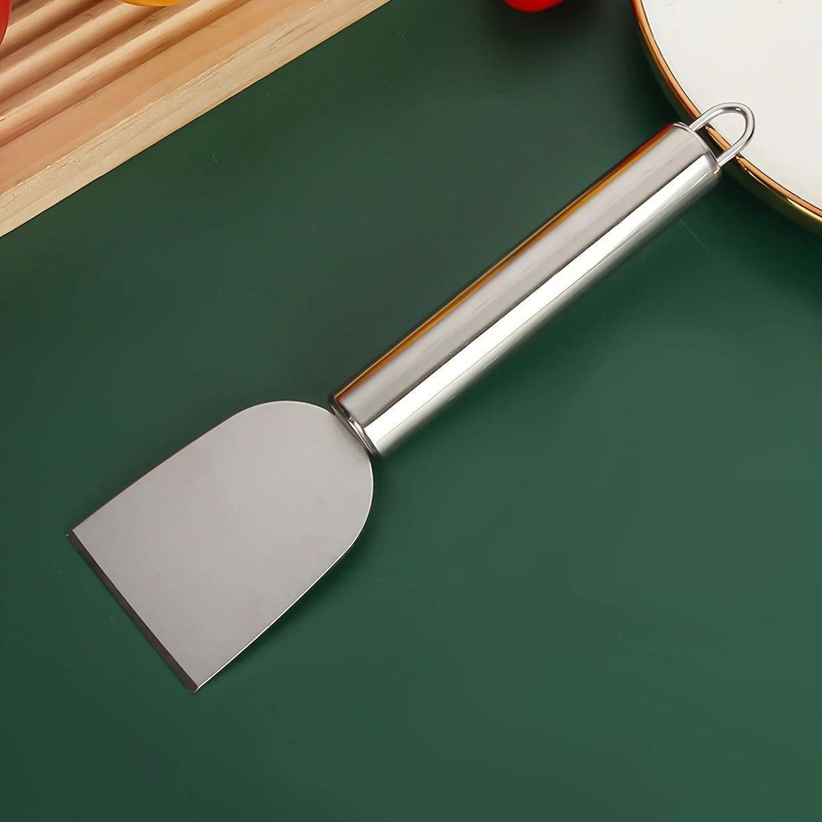 A stainless steel multi-purpose scraper with a handle and blade resting on a dark green surface.