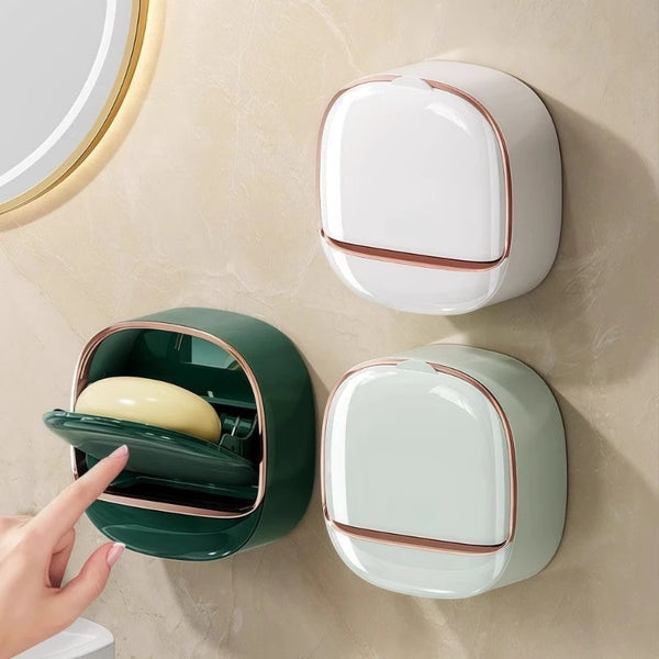 A wall-mounted soap holder in white and green ceramic with a lid, shown against a beige wall.