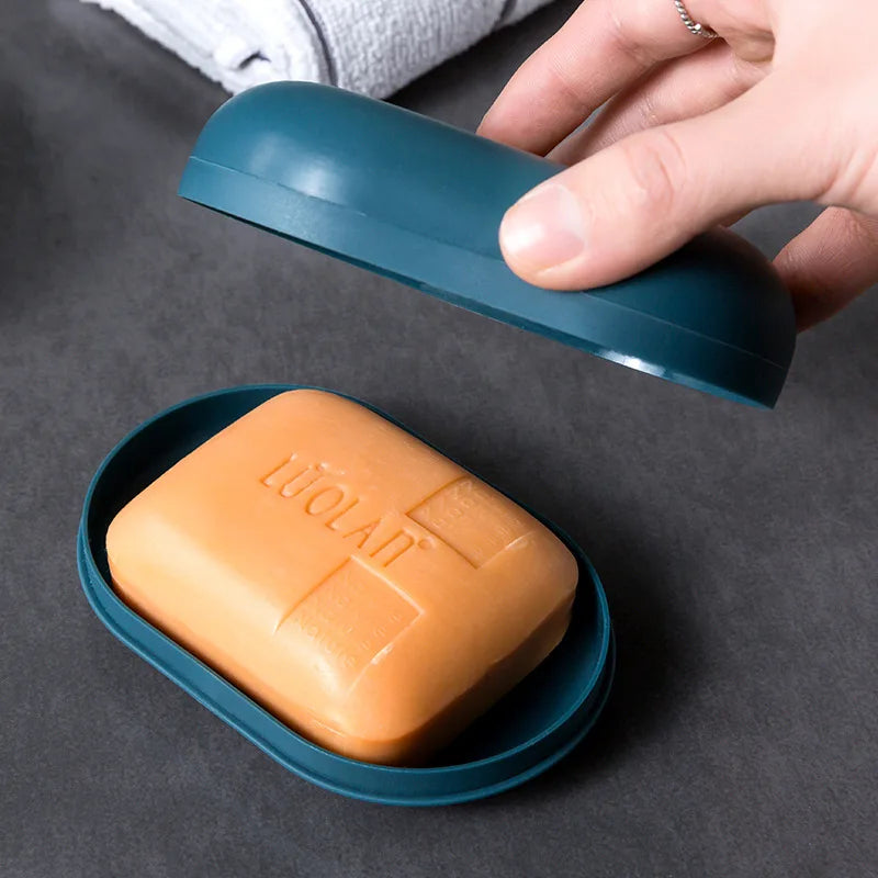 A hand holds a teal portable soap holder with a lid above an orange bar of soap in a blue dish.