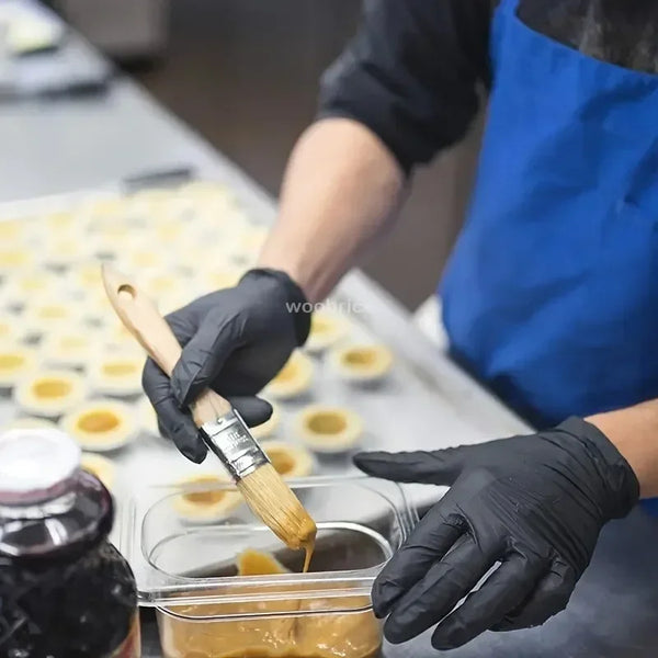 A person wearing black nitrile disposable gloves is using a paintbrush to apply a golden liquid onto a tray of small yellow f