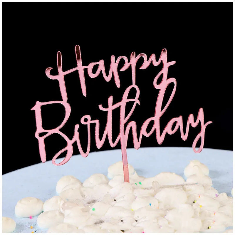 Pink script text spelling Happy Birthday on a cake topper placed on a white surface with white fluffy decorations.