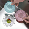 A hand is placing a pink silicone drain strainer into a metal kitchen sink, next to a blue one.