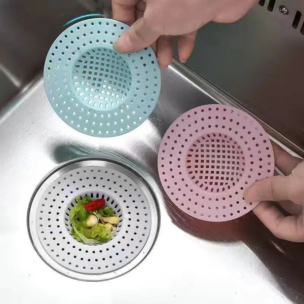 A hand is placing a pink silicone drain strainer into a metal kitchen sink, next to a blue one.