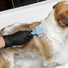 Una persona con guante de nitrilo negro cepilla el pelo de un perro marrón claro y blanco en una bañera llena de espuma de ja