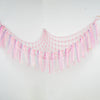 Pink sheer fabric hammock with fringe and tassels hanging against a white wall.