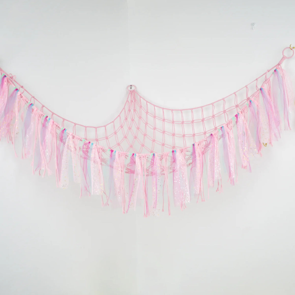 Pink sheer fabric hammock with fringe and tassels hanging against a white wall.