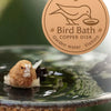 A small brown bird is drinking water from a copper disk bird bath in a garden setting.