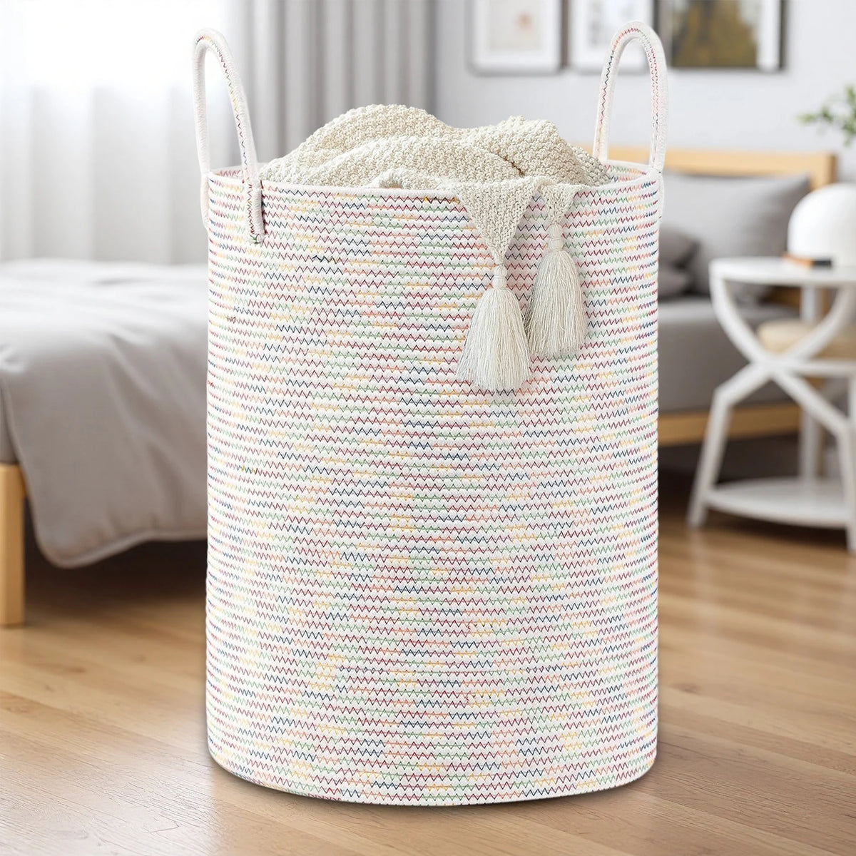 A large woven storage hamper in a light pink and cream striped pattern with tassels, sitting on a wooden floor.